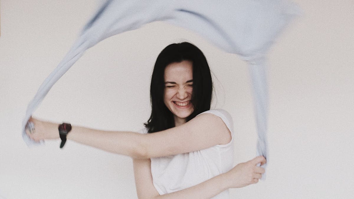 Happy woman with eyes closed joyfully waving a scarf indoors against a simple background.